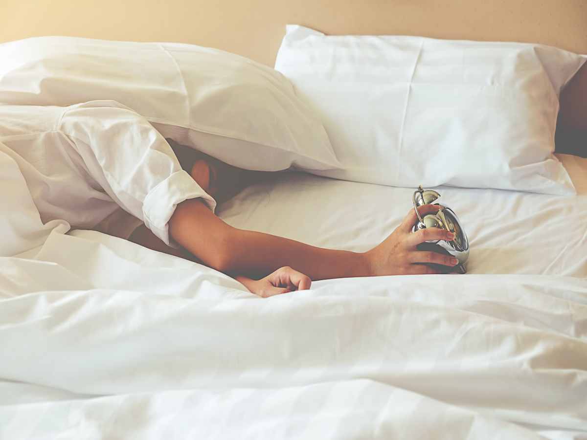 persona con la testa sotto il cuscino prova a spegnere la sveglia